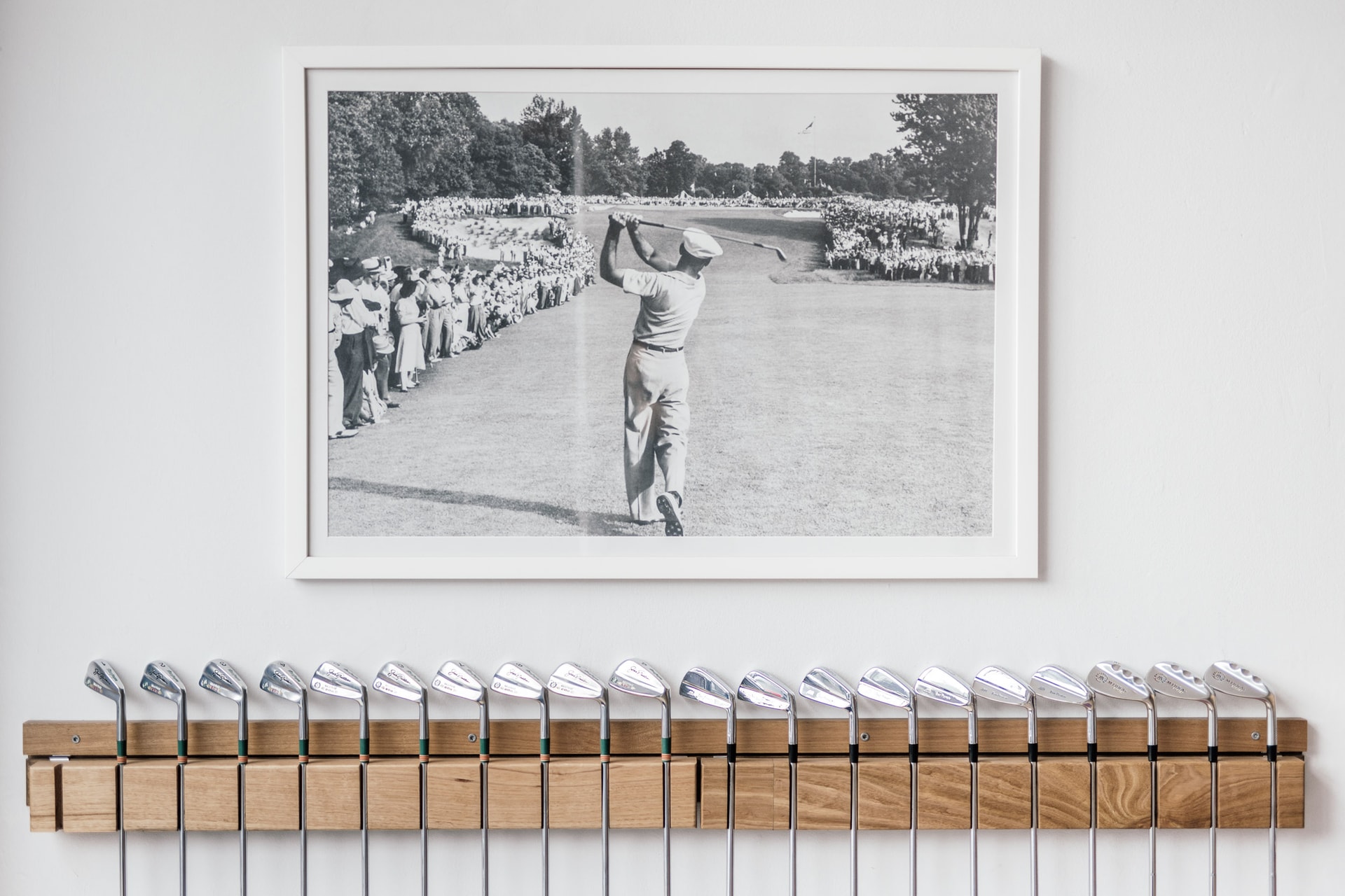 A framed black and white photograph of a golfer finishing his swing before a lined crowd, above a long rack of irons