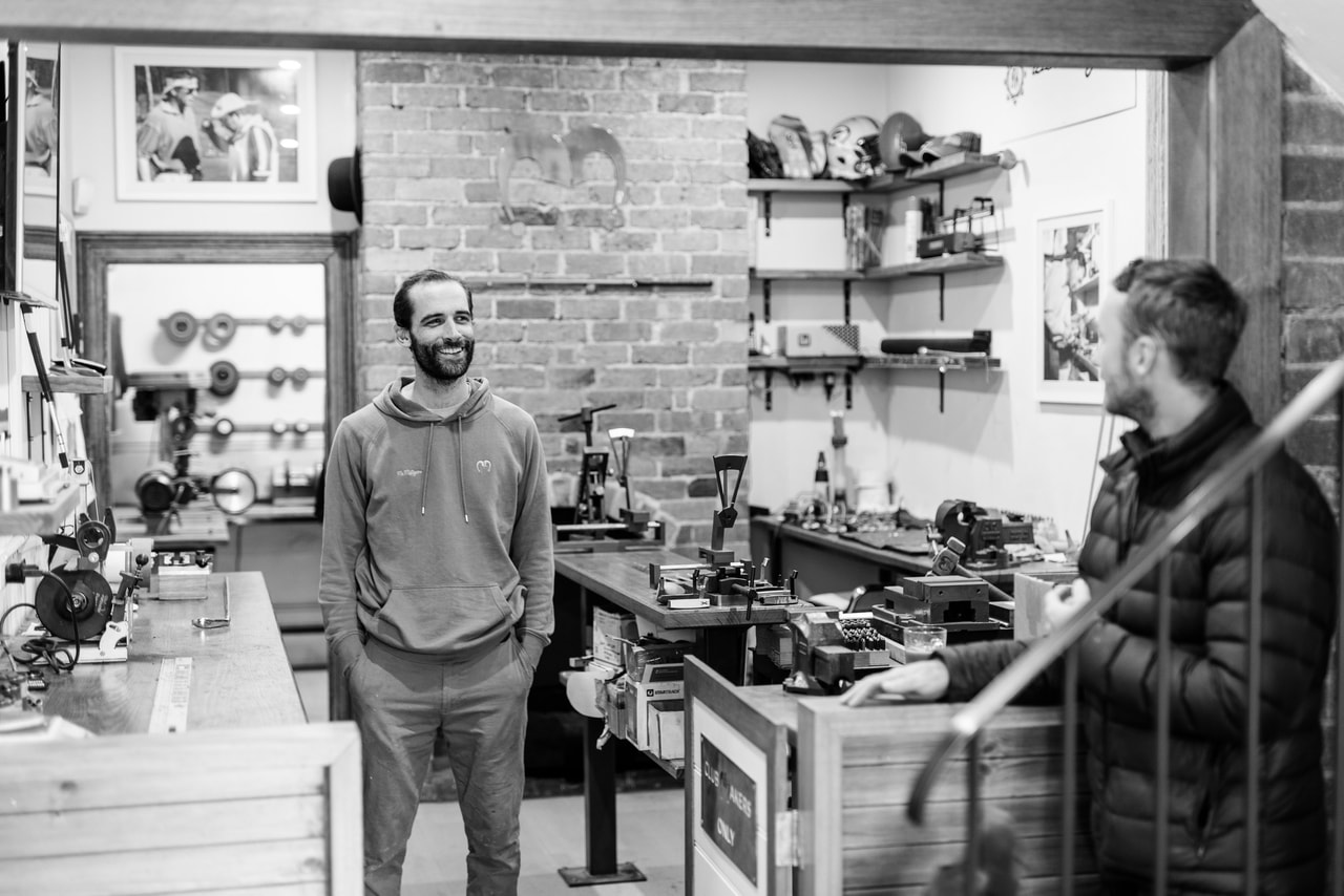 A smiling man in a hoodie stands among workbenches and vices, another man in a puffer jacket in the foreground, in black and white
