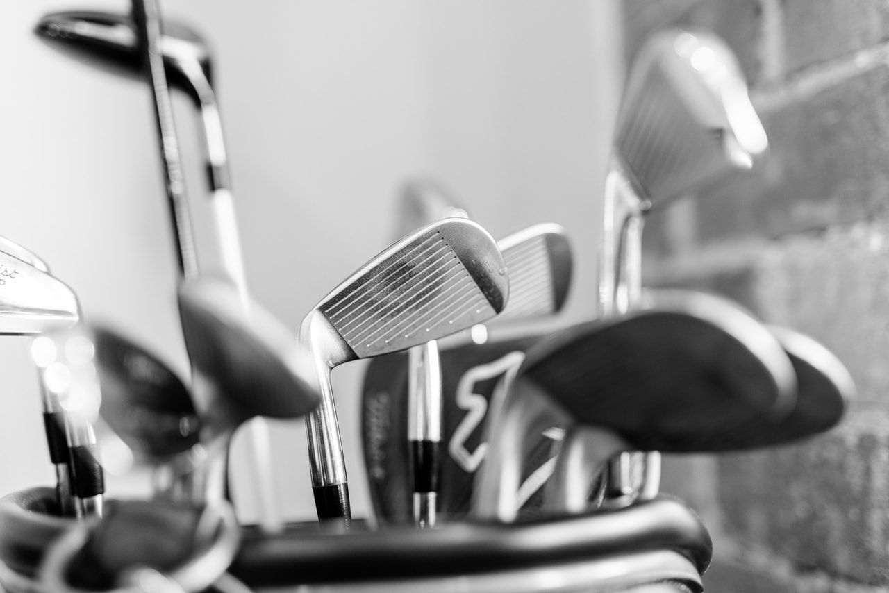 Irons and headcovered woods standing in a golf bag against a brick wall, in black and white