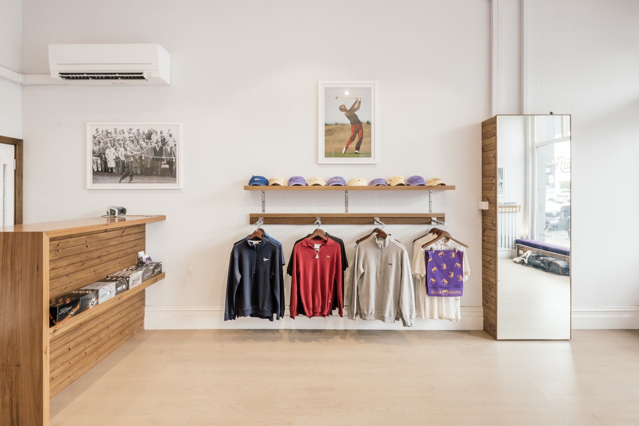 A shop floor with a timber counter, jumpers and shirts on a rail, caps on a shelf and framed golf photographs on white walls