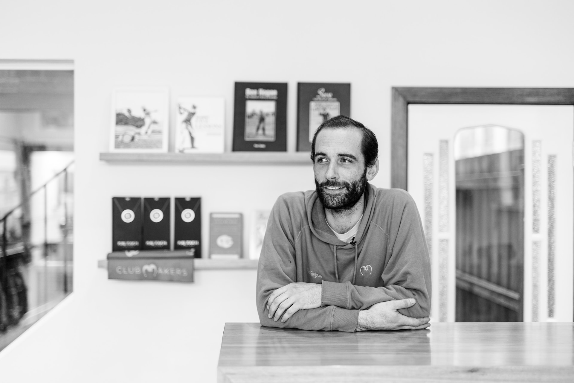 A bearded man in a hoodie leans, arms folded, on a timber counter, shelves of golf books and boxes behind, in black and white