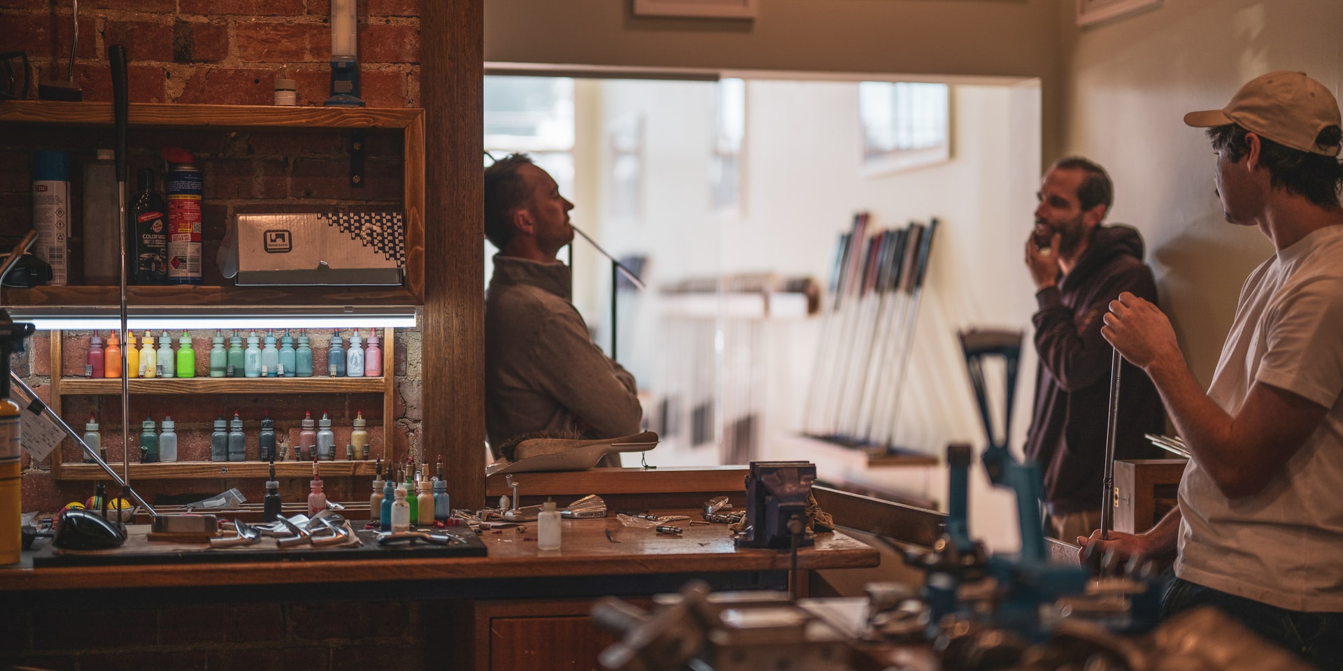 Three men talk in a workshop with brick walls, shelves of paint bottles and clubs in a rack behind