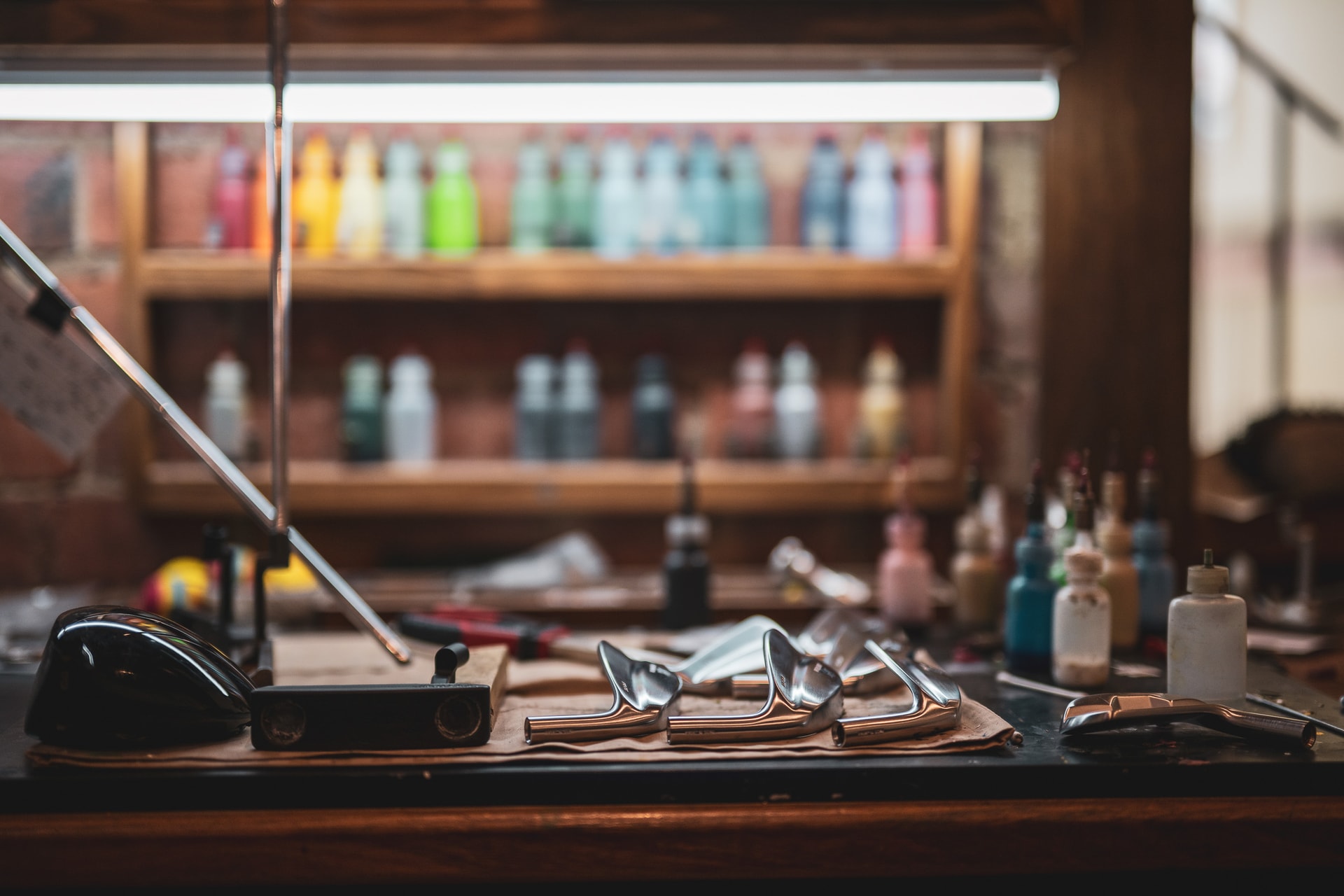 A workbench of putter and iron heads, shelves of coloured paint bottles lit behind