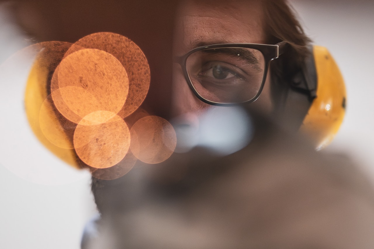 A man in glasses and yellow ear muffs looks through a glow of sparks