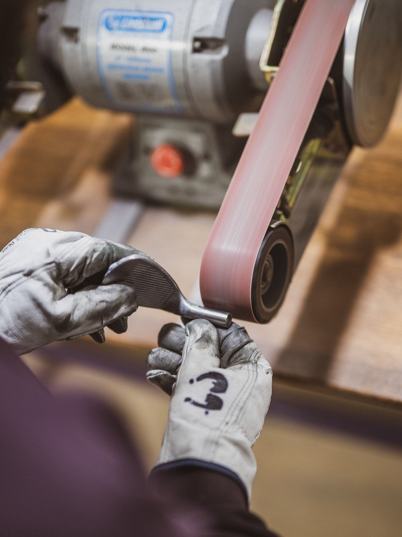 Gloved hands hold a wedge head against a belt sander, one glove marked in black pen