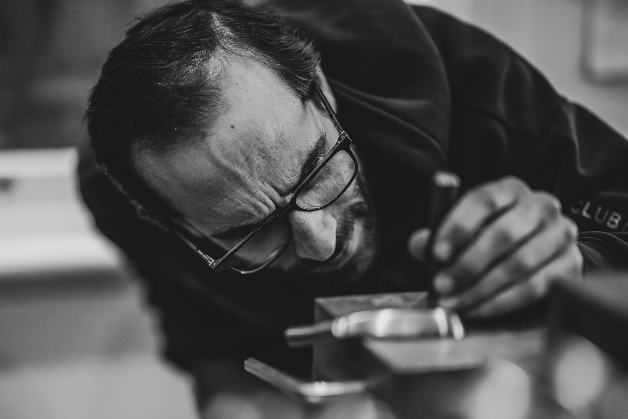 A man in glasses bends close over a club head in a vice, working it with a small hand tool, in black and white