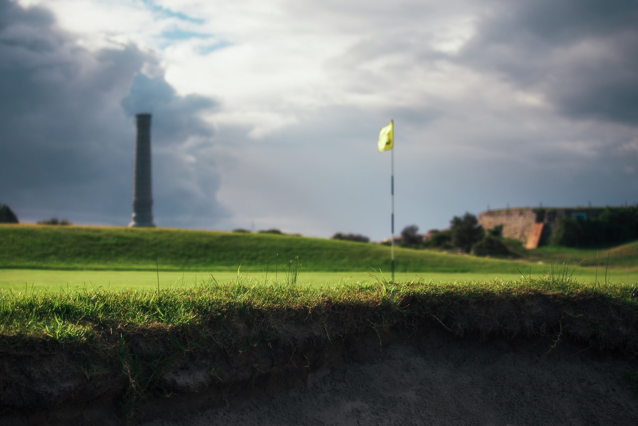 From a bunker’s edge, a yellow flag on the green, the vent stack beyond under dark storm cloud