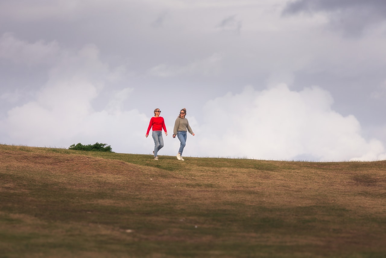 Two women in jeans, one in a red jumper, stroll across a fairway under billowing cloud