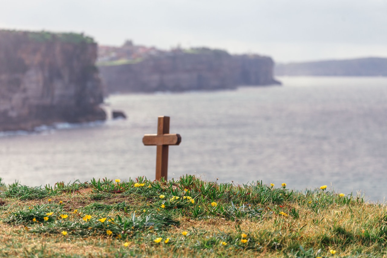 A small wooden cross among yellow wildflowers on the clifftop, sandstone cliffs and sea beyond