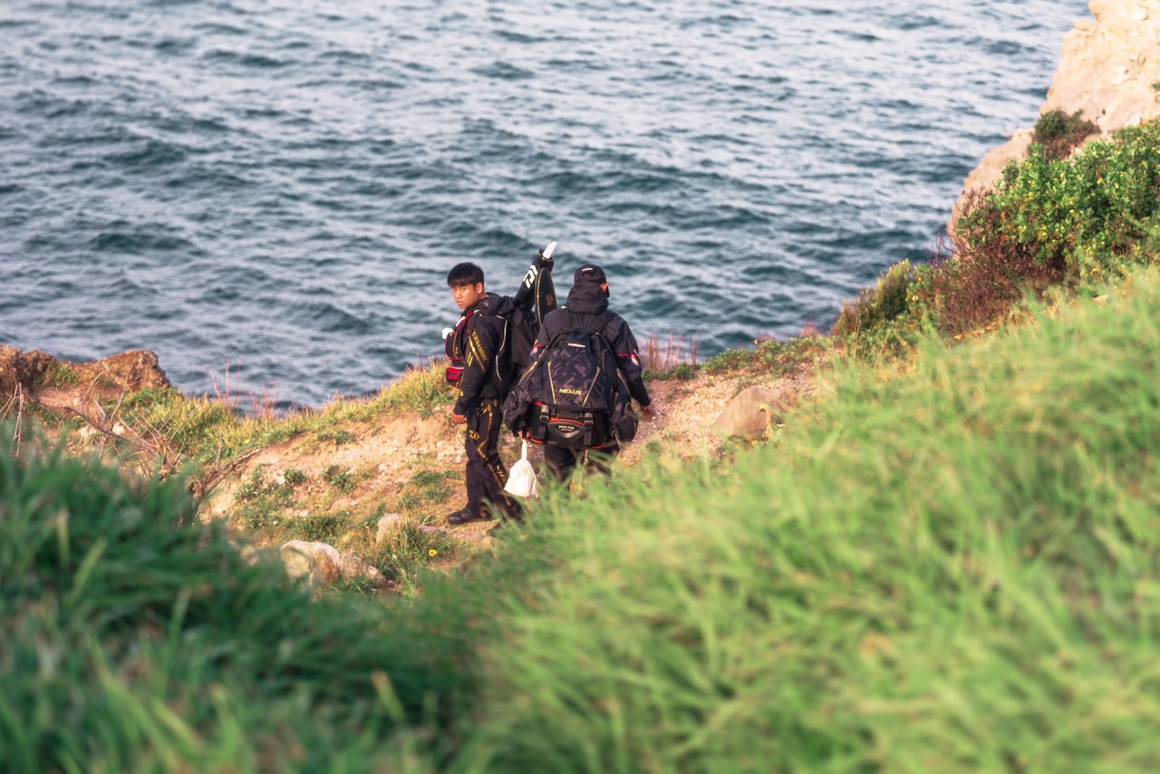 Two men in black fishing suits and packs pick their way down a cliff path towards the sea