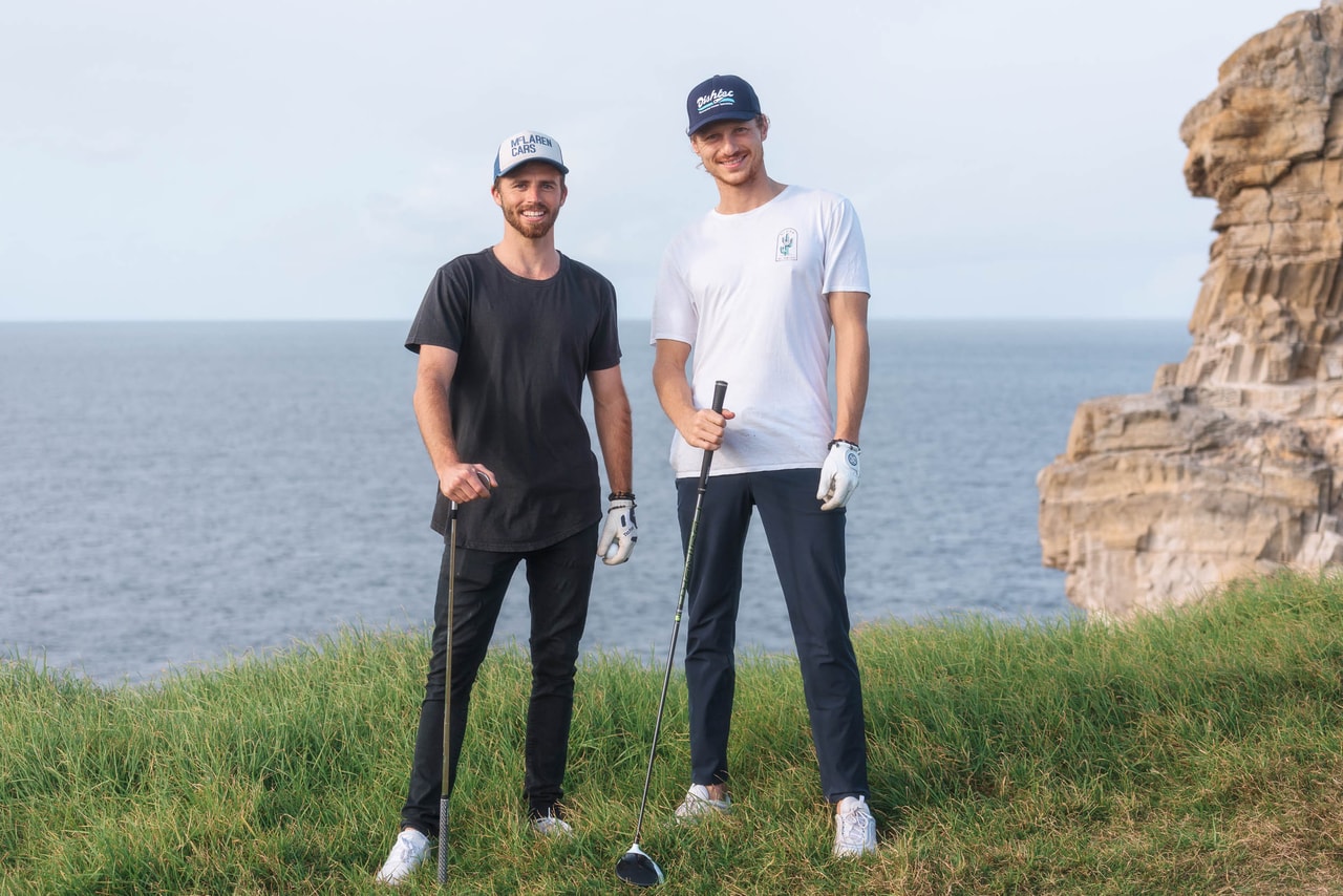 Two young men in caps, one in black, one in white, hold drivers on a seaside clifftop