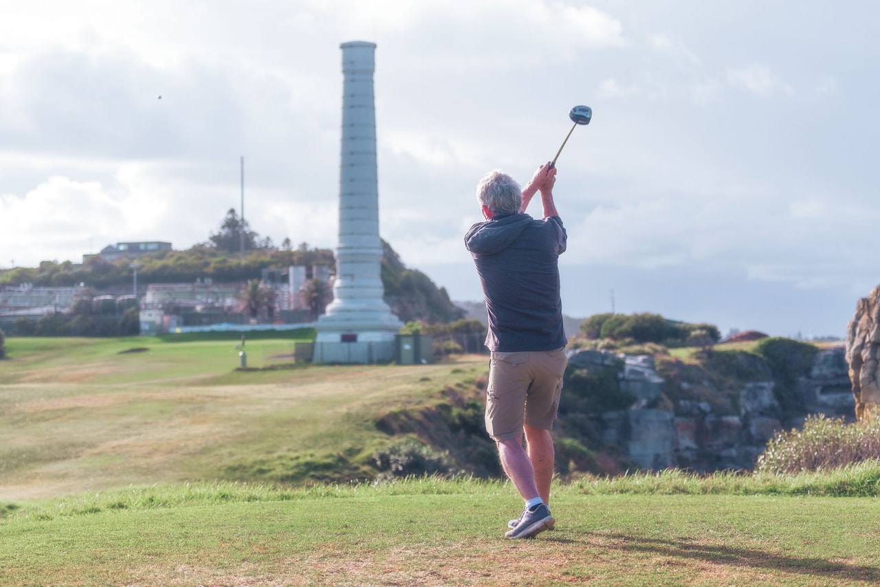 A grey-haired golfer in a hoodie and shorts follows through on the tee, the tall vent stack ahead