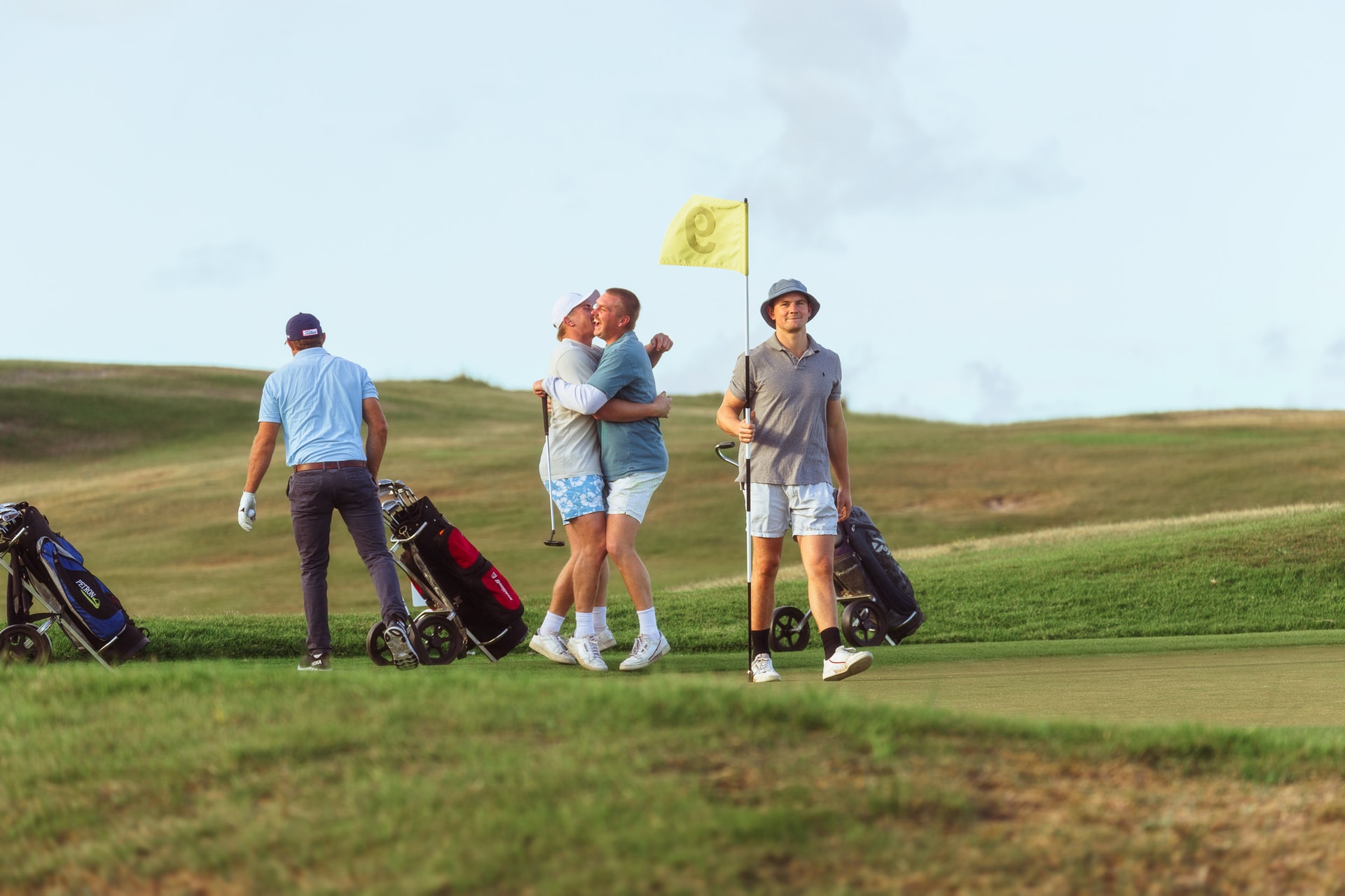Four golfers on a green, two hugging as another holds the yellow flag, trolleys beside them