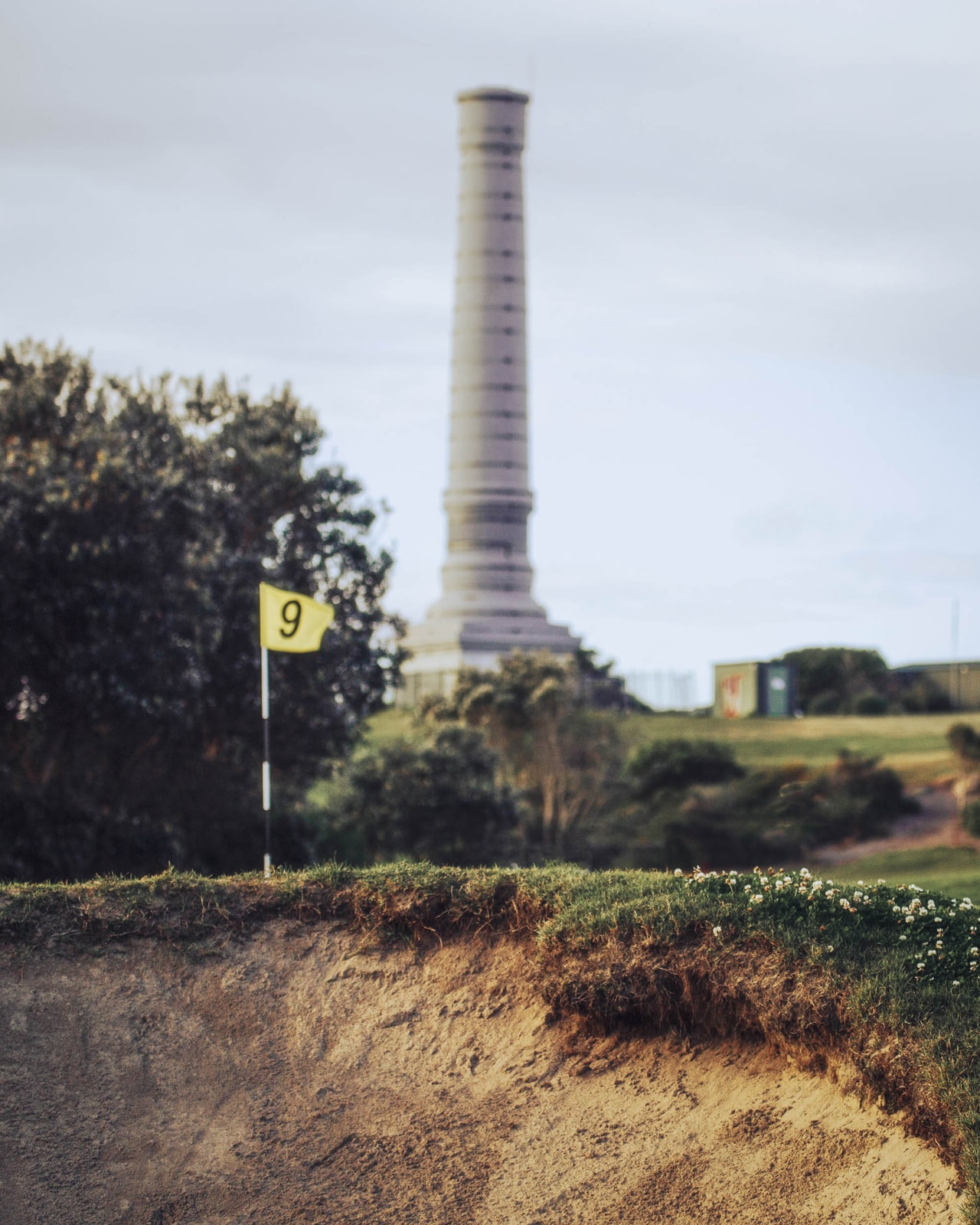 A yellow flag at a green’s edge above a sandy bank, a tall stone vent stack behind