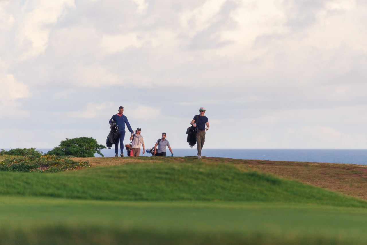 Four golfers with their bags come over a grassy rise, the sea behind under soft cloud