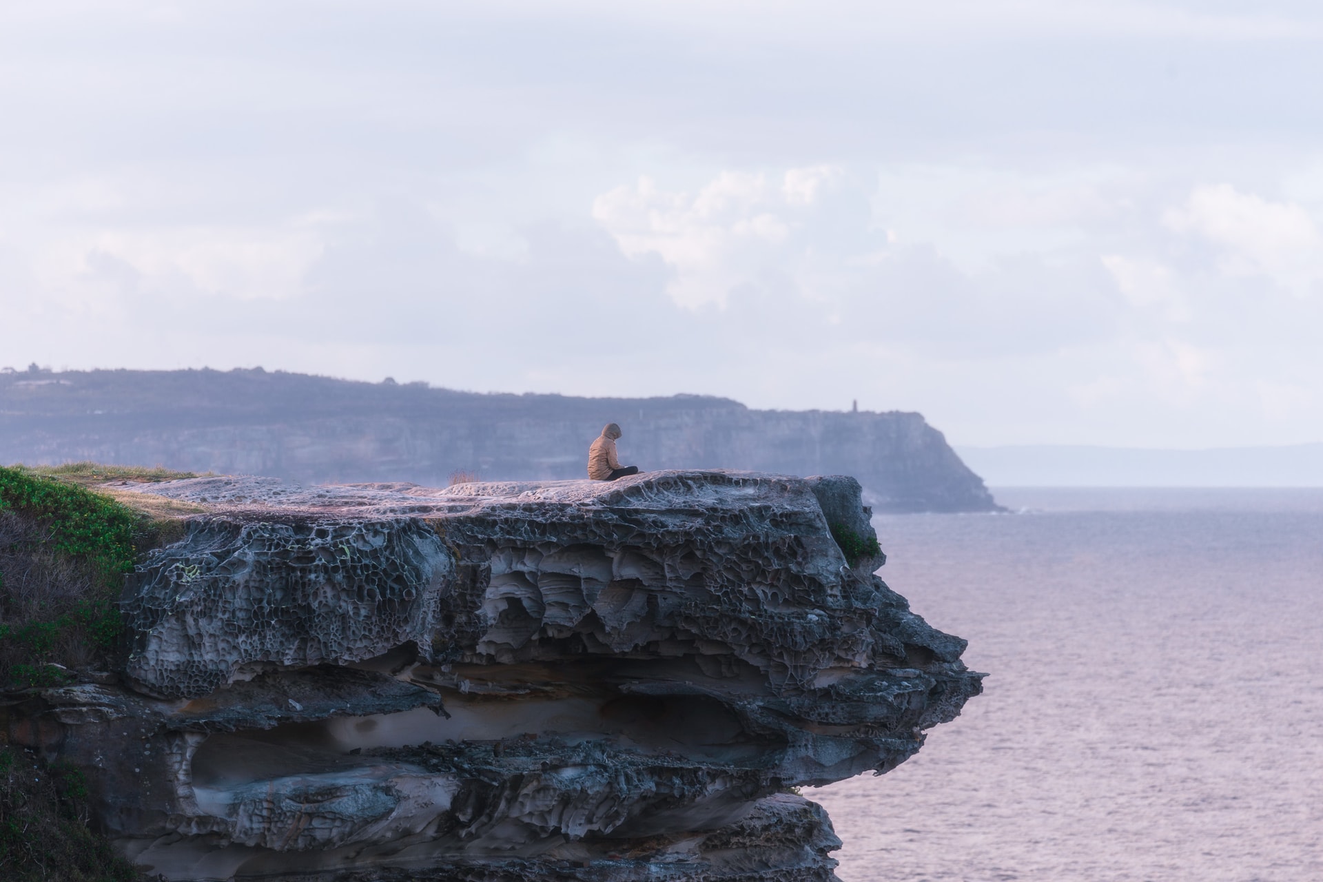 A lone figure sits on the edge of a weathered sandstone ledge above the sea, a headland beyond