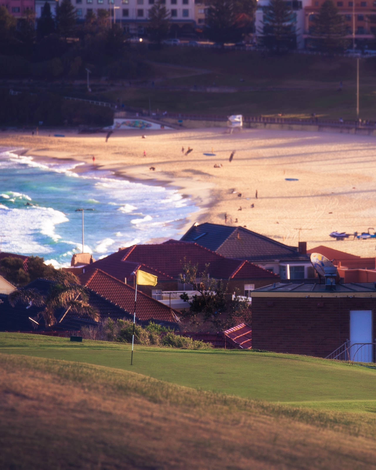 A green and its yellow flag above red-tiled roofs, the surf and sand of Bondi Beach beyond