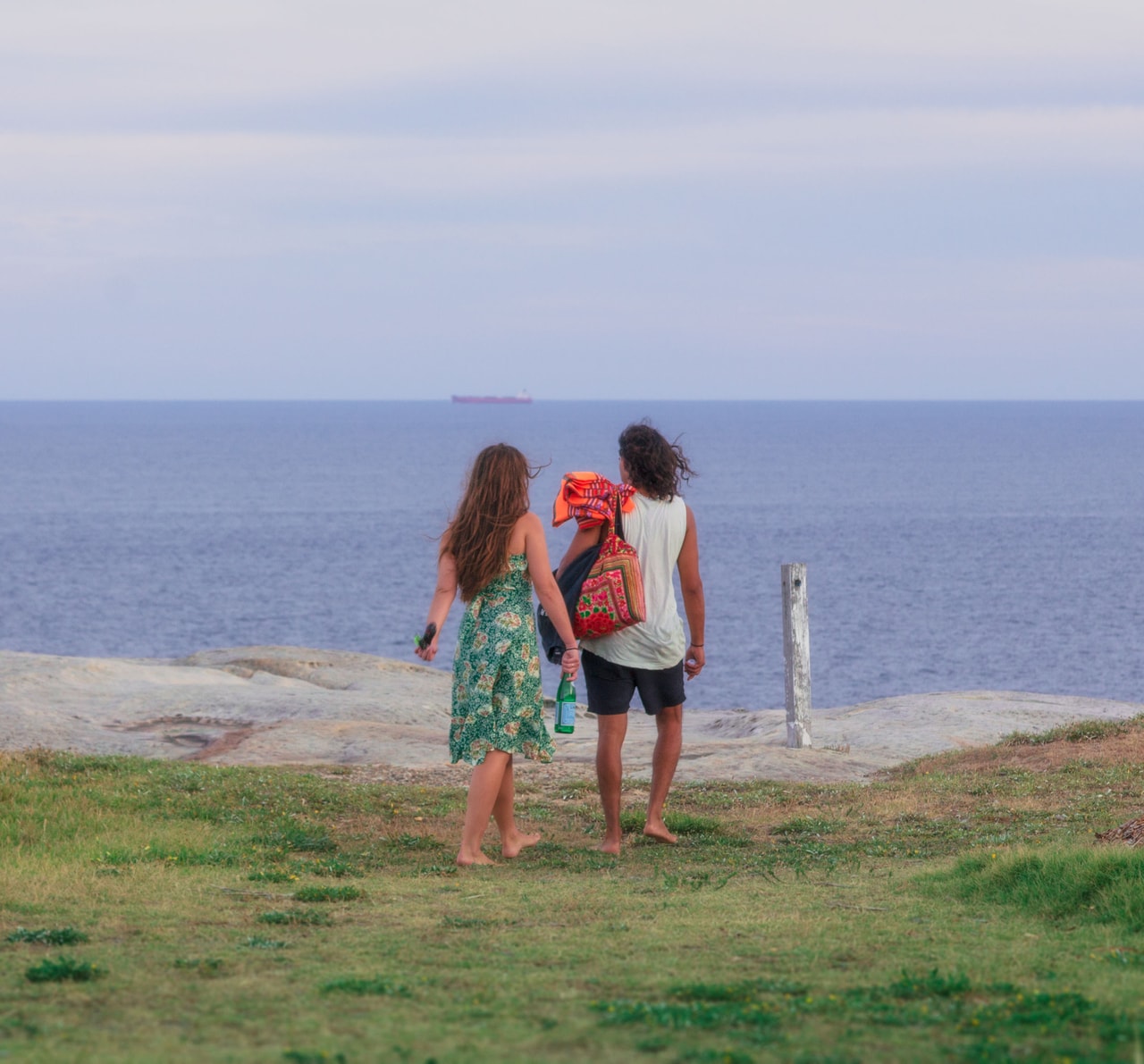 A barefoot couple, one in a green floral dress, walk towards the rock shelf and the sea