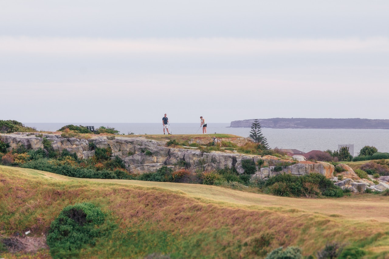 Two golfers on the clifftop above sandstone ledges and scrub, the sea and a long headland beyond