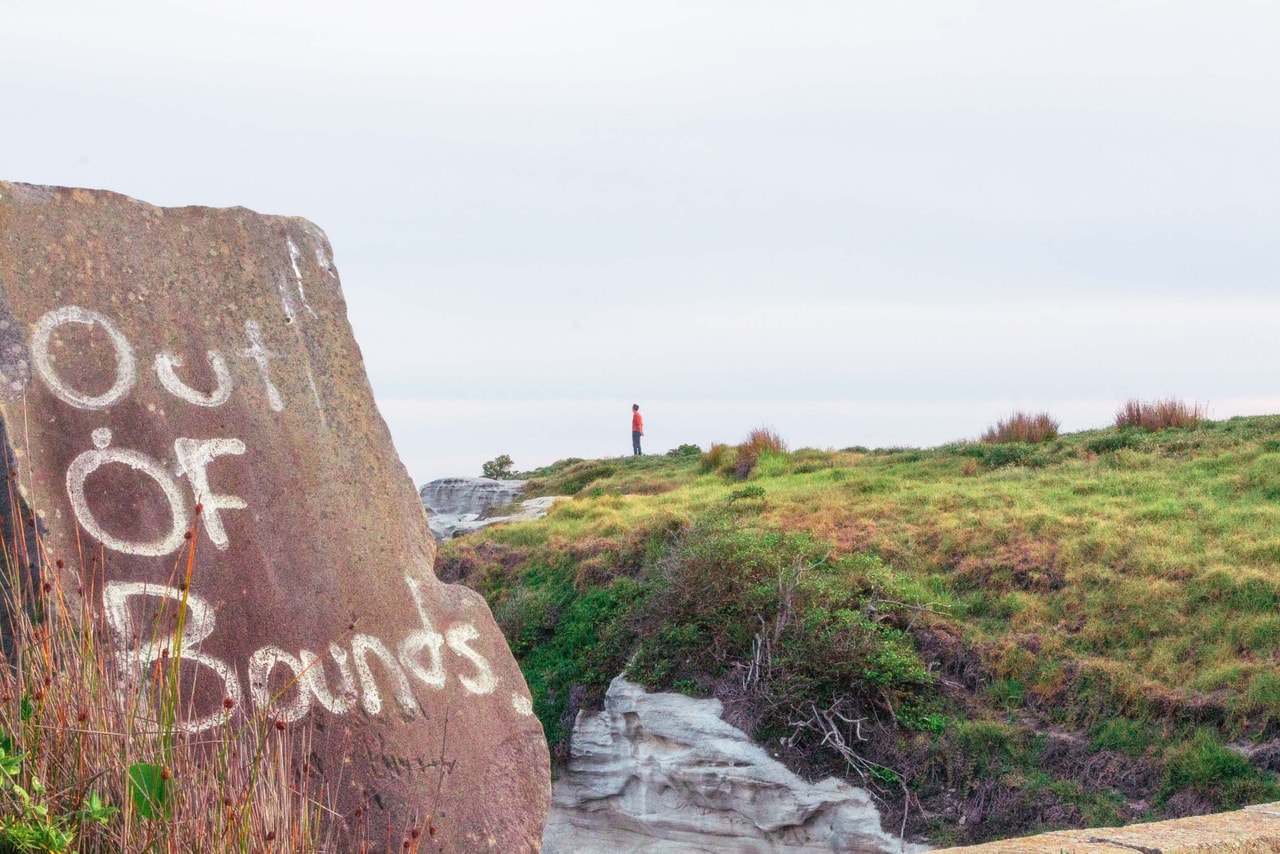 A boulder painted ‘Out of Bounds’ in white, a figure in red on the grassy clifftop beyond