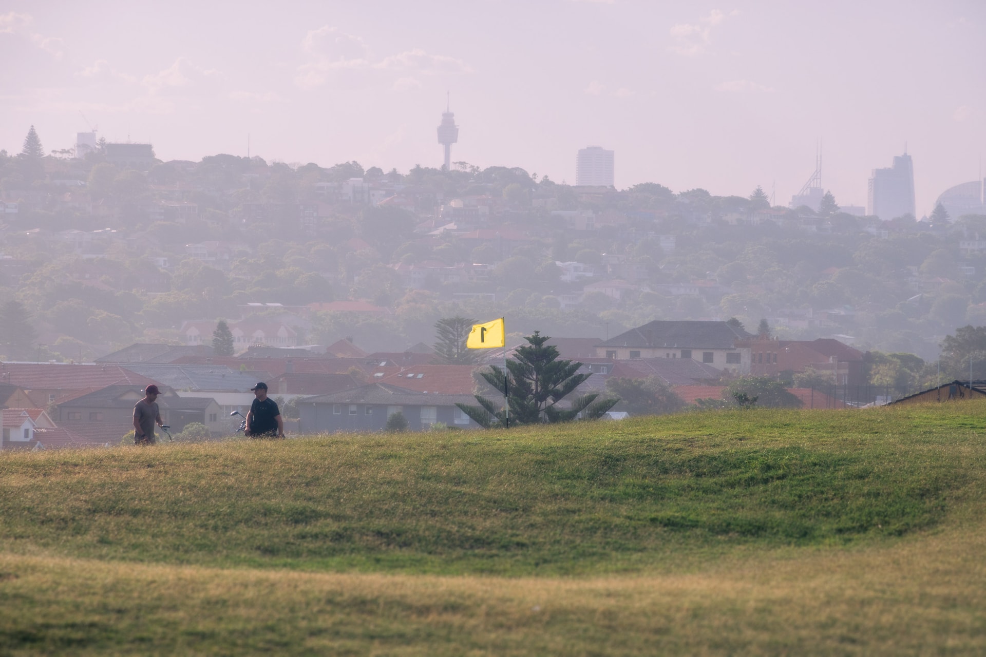 Two golfers on a grassy rise by a yellow flag, rooftops and the hazy Sydney skyline beyond