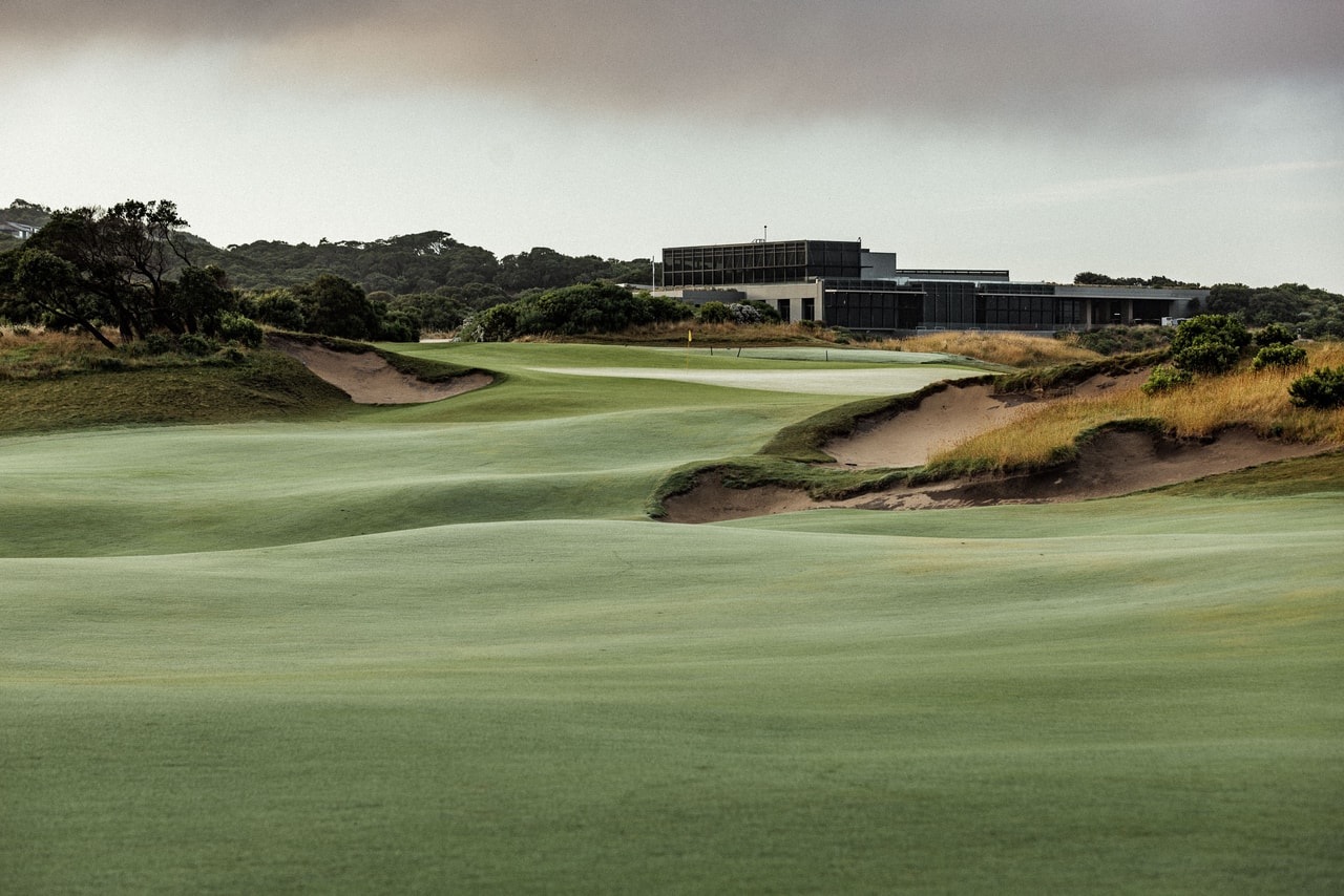 A rumpled fairway running between ragged bunkers to a green, a long modern building on the rise beyond