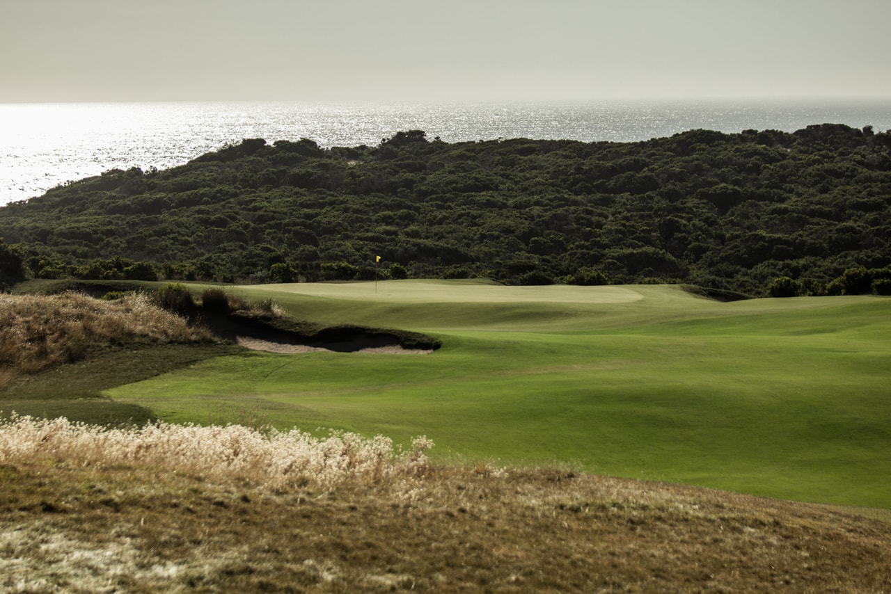 A green beyond a bunker and pale tussock grass, dense coastal scrub and a glittering sea behind