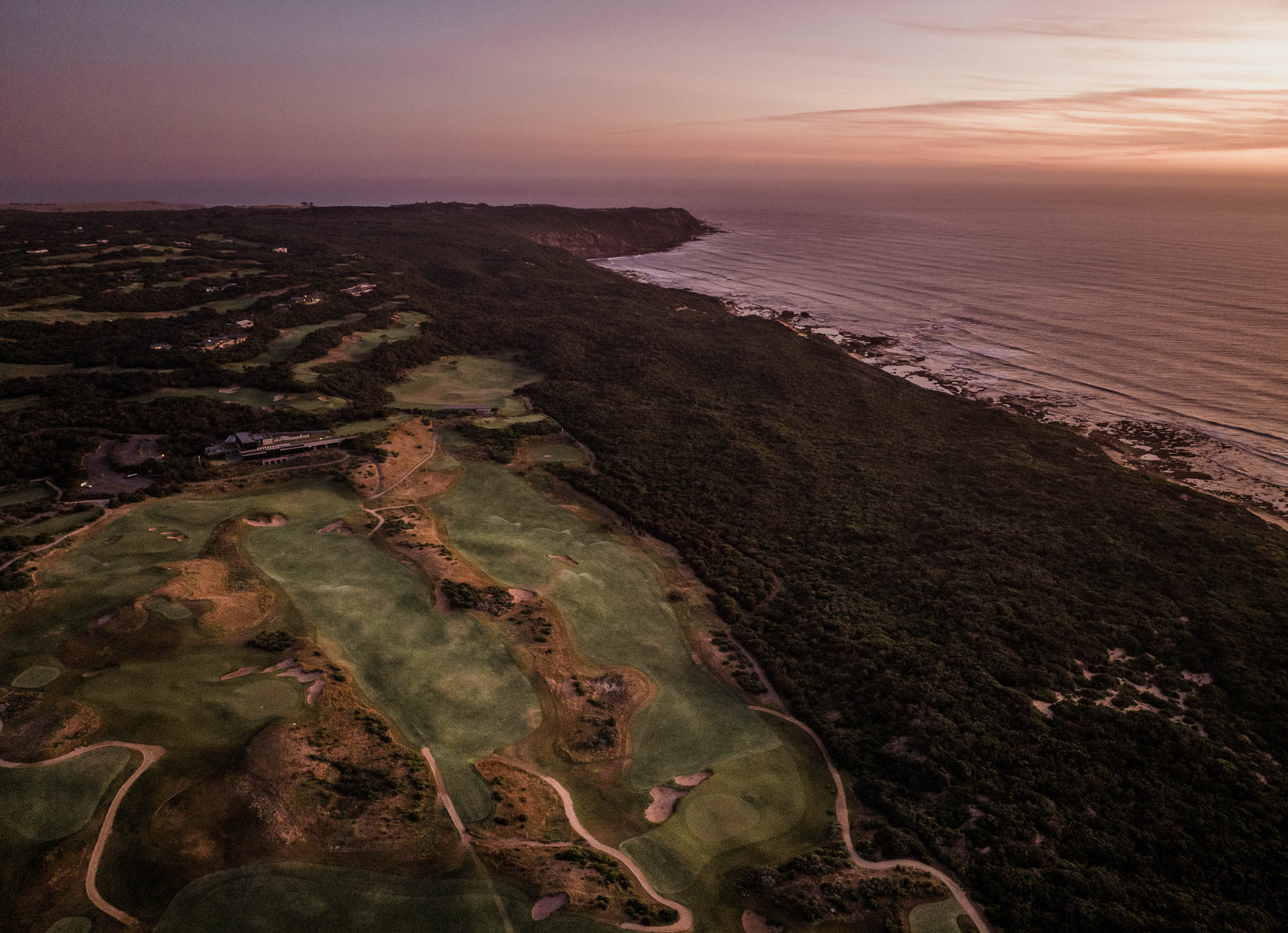 The course from the air at dusk, fairways in coastal scrub along the clifftops, surf below a pink sky