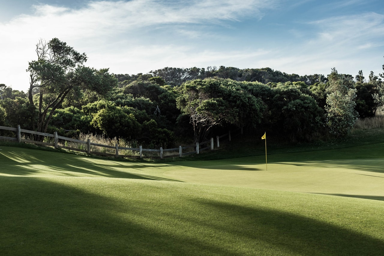 A green and its flag in long shadow, a post-and-rail fence and dense coastal trees behind