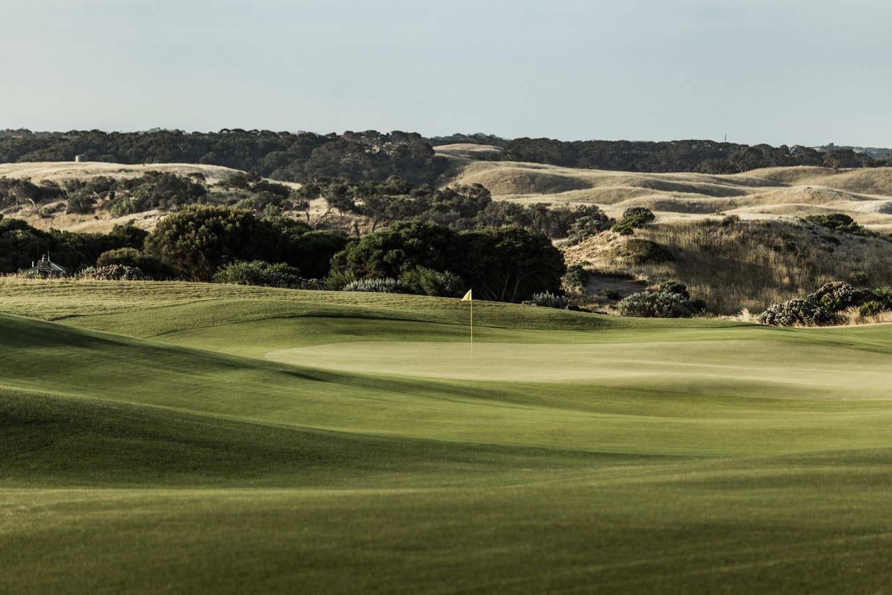 A rolling green and its flag before dense coastal scrub and sandy dunes