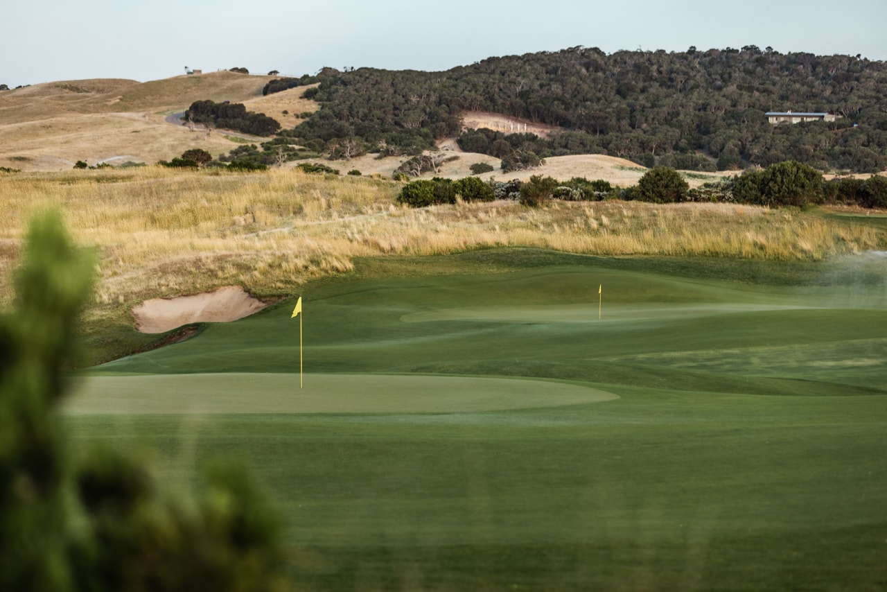 Two greens and their flags beyond a bunker in golden grass, wooded hills behind