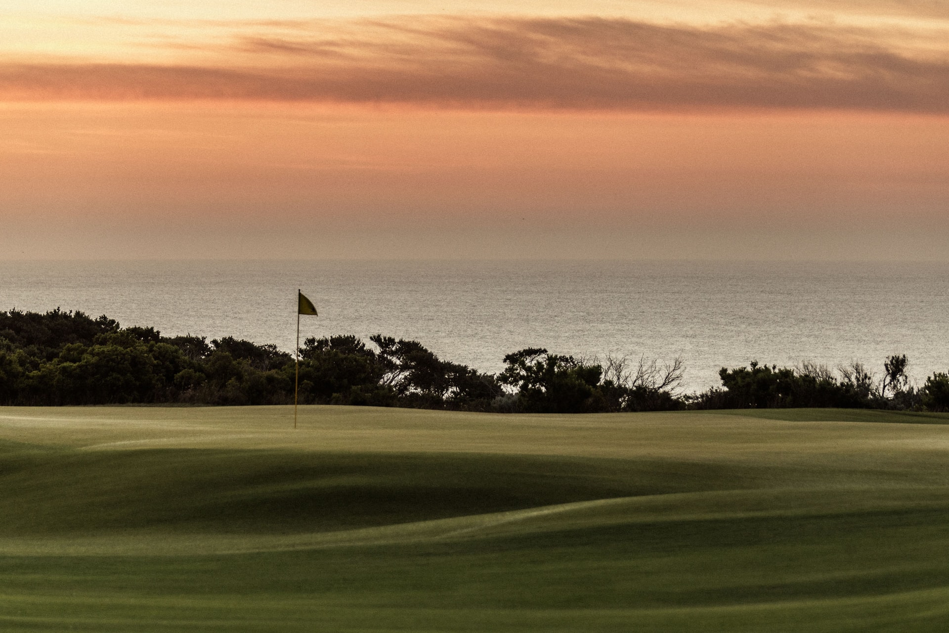 A flag on a green against the sea at dusk, dark scrub between and an orange sky above