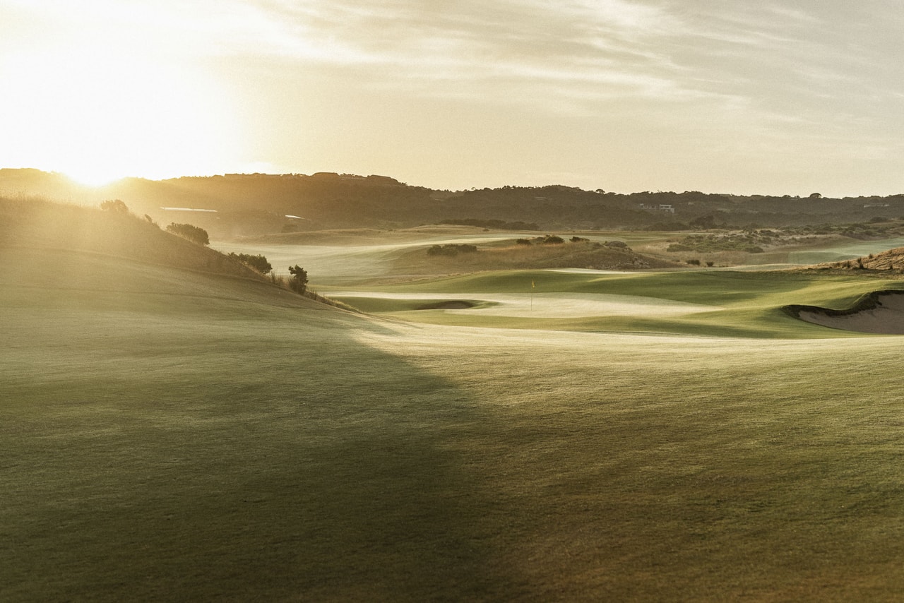 Low sun spilling over rolling fairways and dunes, a distant flag and bunkers in soft haze