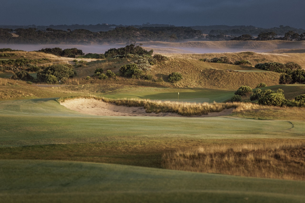 A grass-fringed bunker in low sun, a green and flag beyond, scrubby dunes and mist under a dark sky