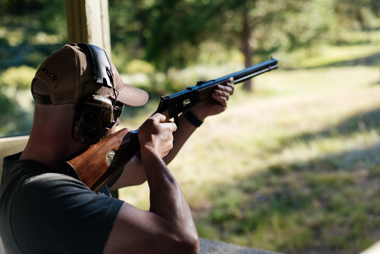 A man in a cap and ear defenders aims a wood-stocked rifle from a shelter, sunlit grass and pines beyond