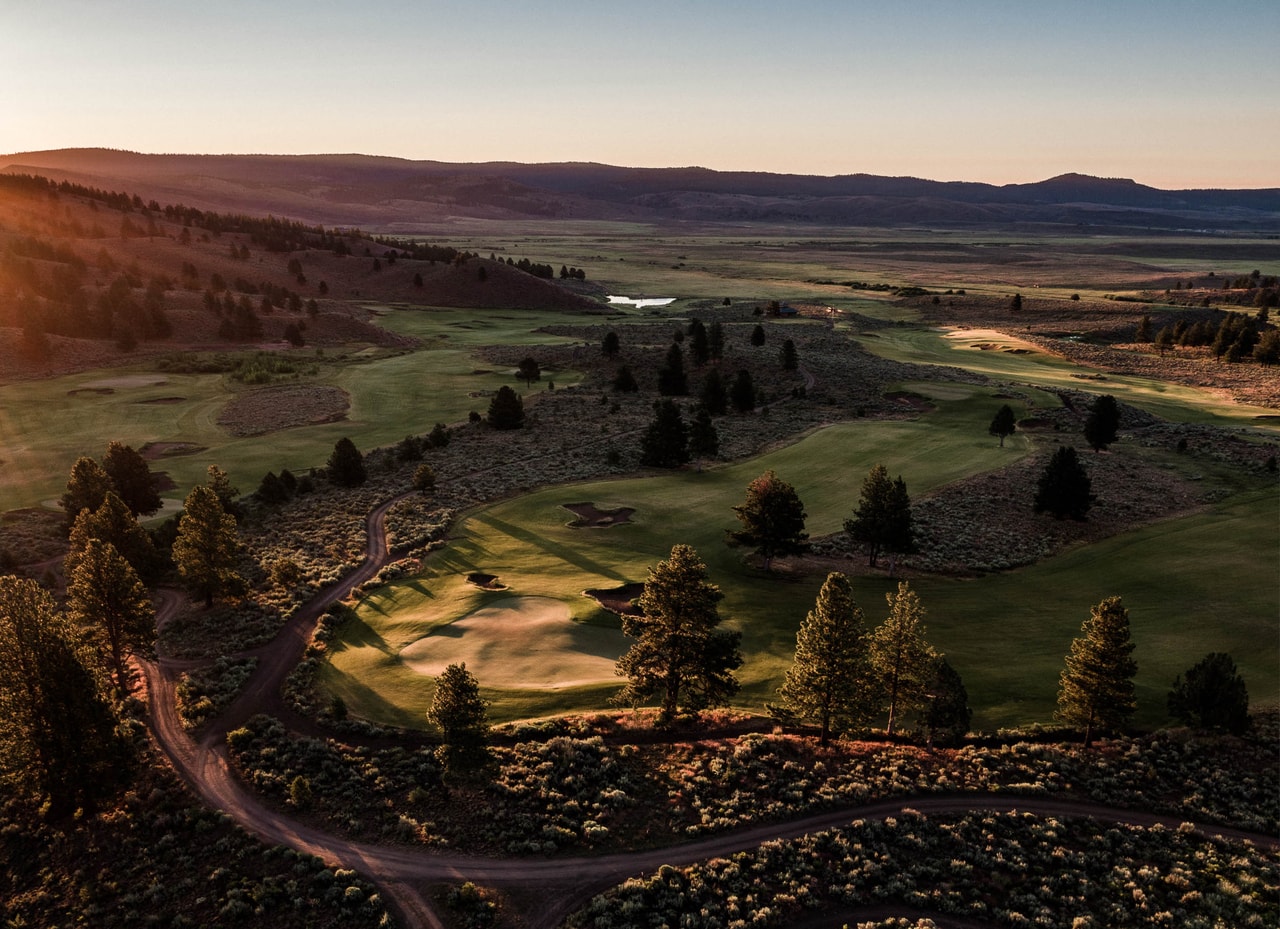 Bunkered fairways among pines and sage from the air in warm low light, a cart path curving through, the valley and hills beyond