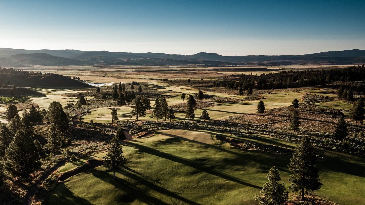 Fairways among pines casting long shadows, seen from the air, a broad valley and distant ranges beyond in low sun