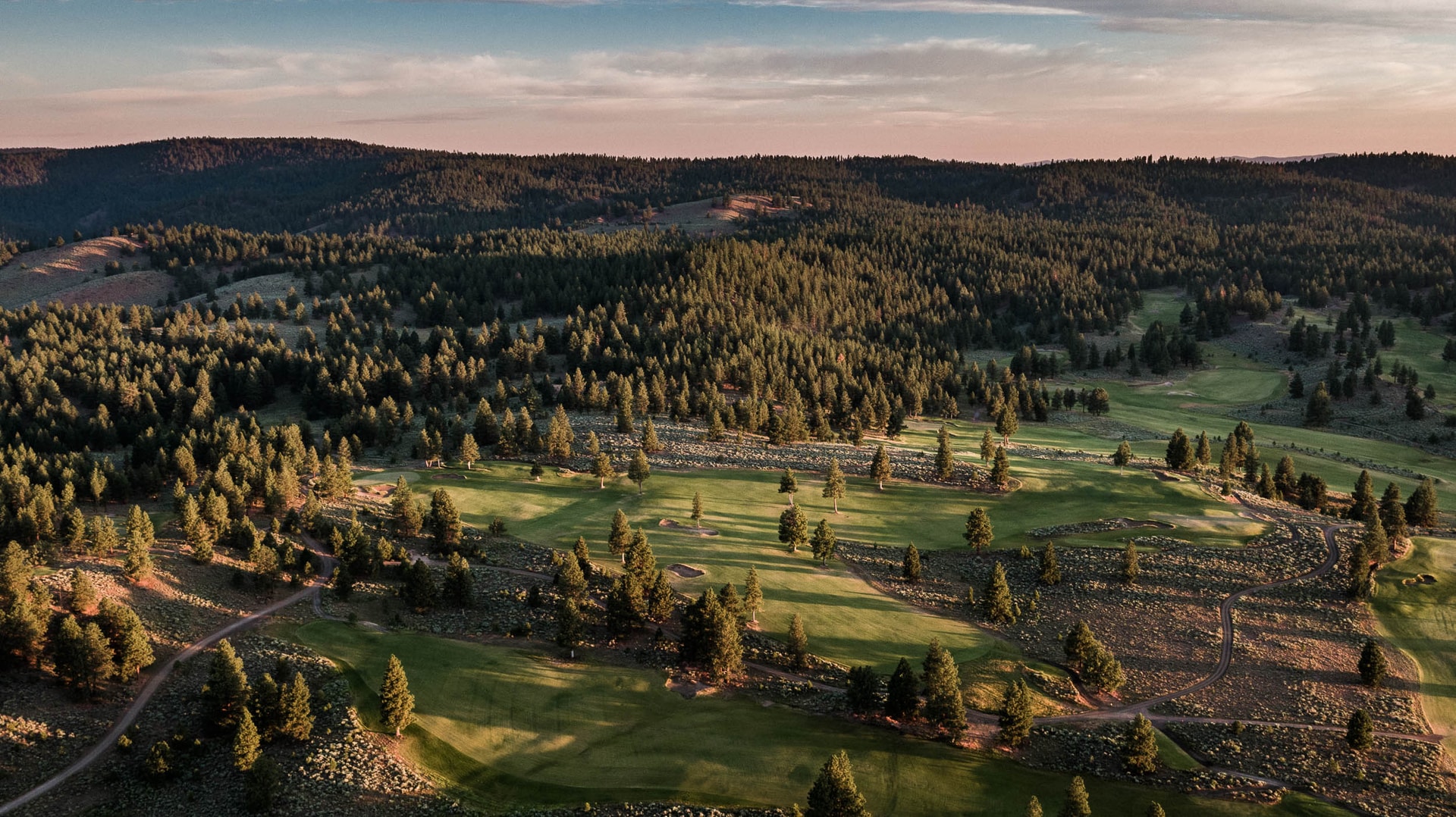 Fairways threading through sage and scattered pines below forested hills, seen from the air under a pink-tinged sky