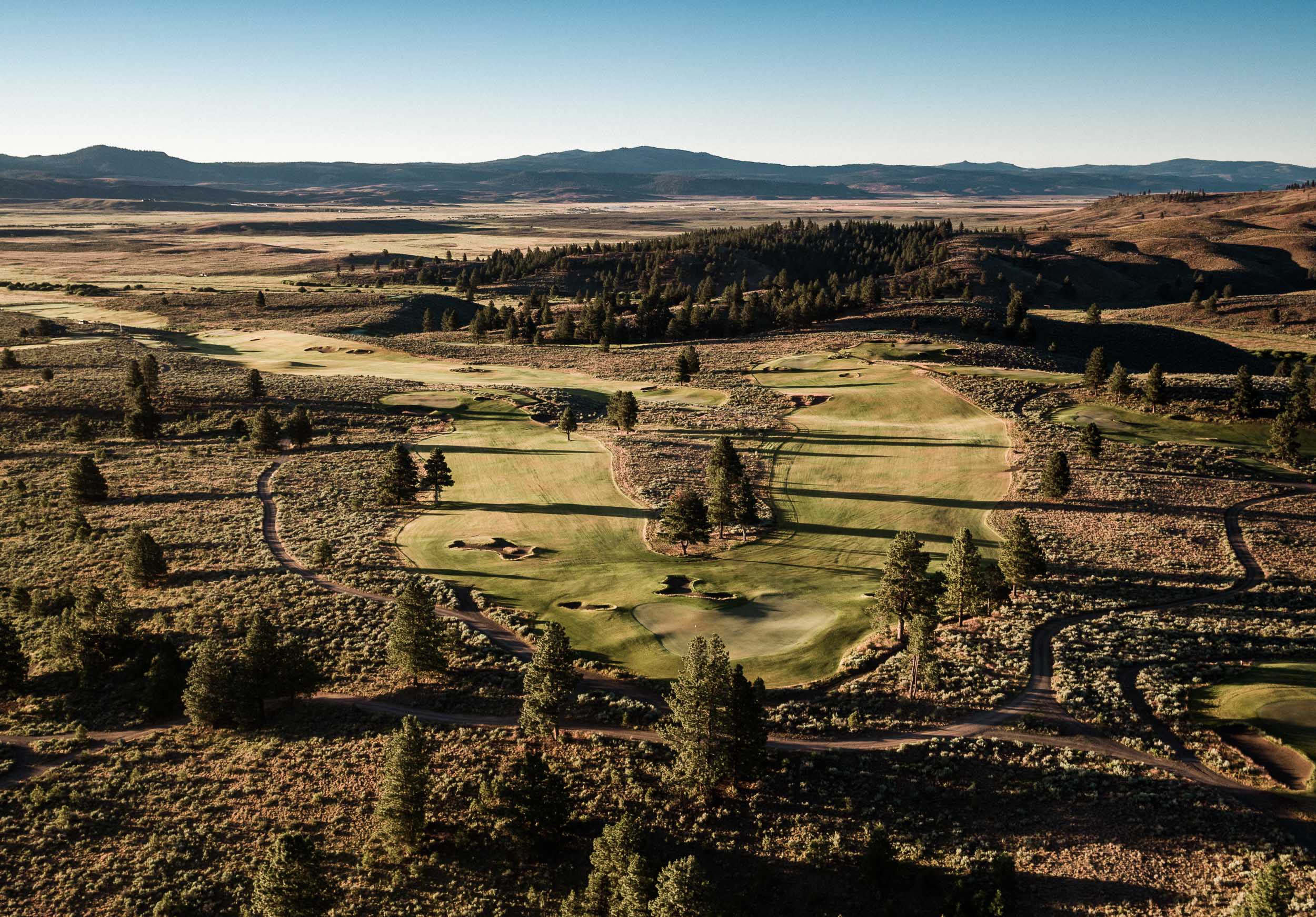 A fairway and bunkered green among sage and pines from above, cart paths winding, open plains and distant ranges beyond