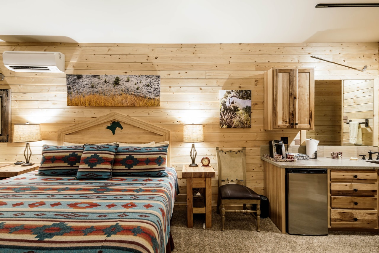 A pine-panelled guest room: a bed in patterned wool blankets, lamps lit either side, a kitchenette with a bar fridge