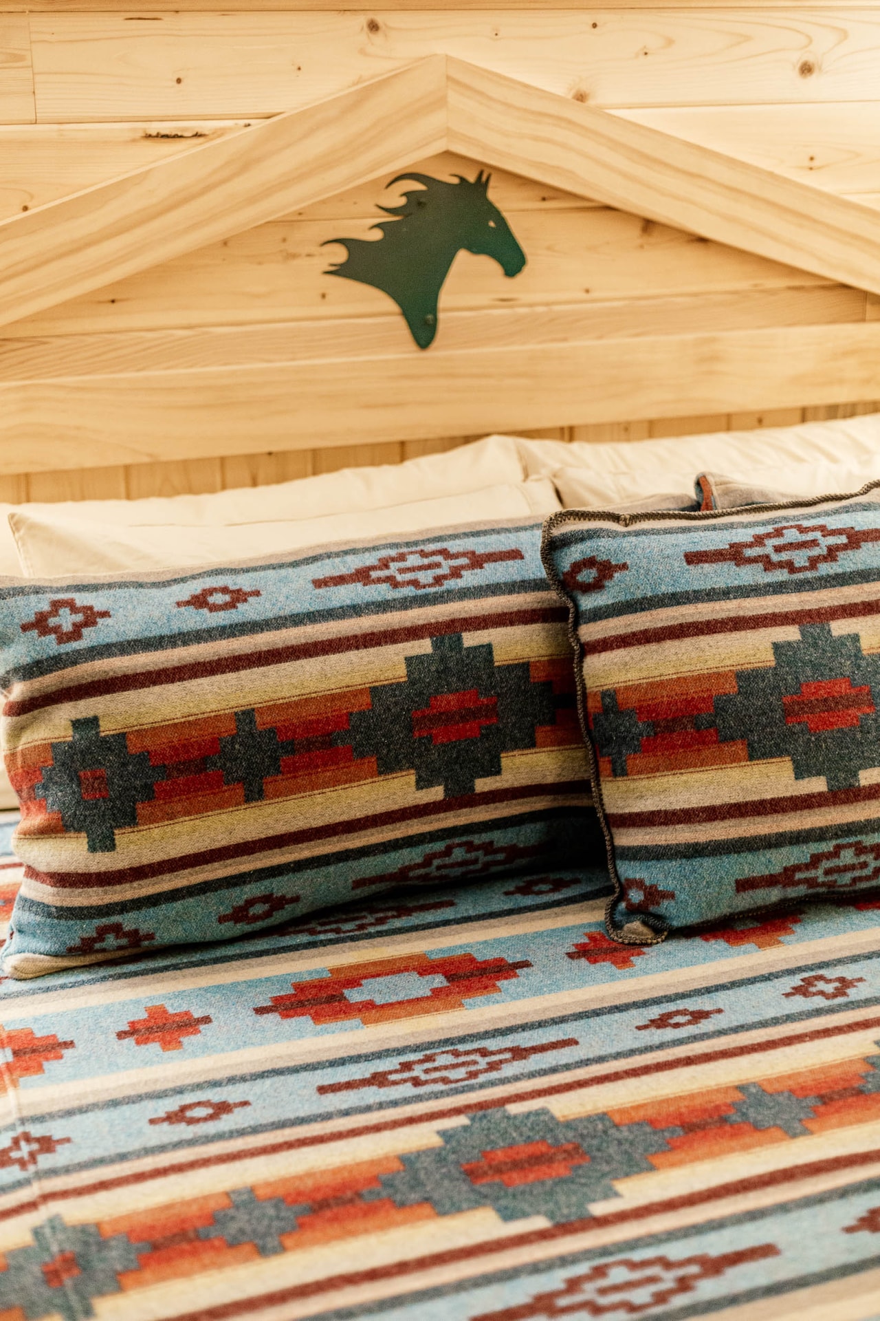 Pillows and a bedspread in a turquoise, red and grey geometric pattern below a pine headboard with a cut-metal horse’s head