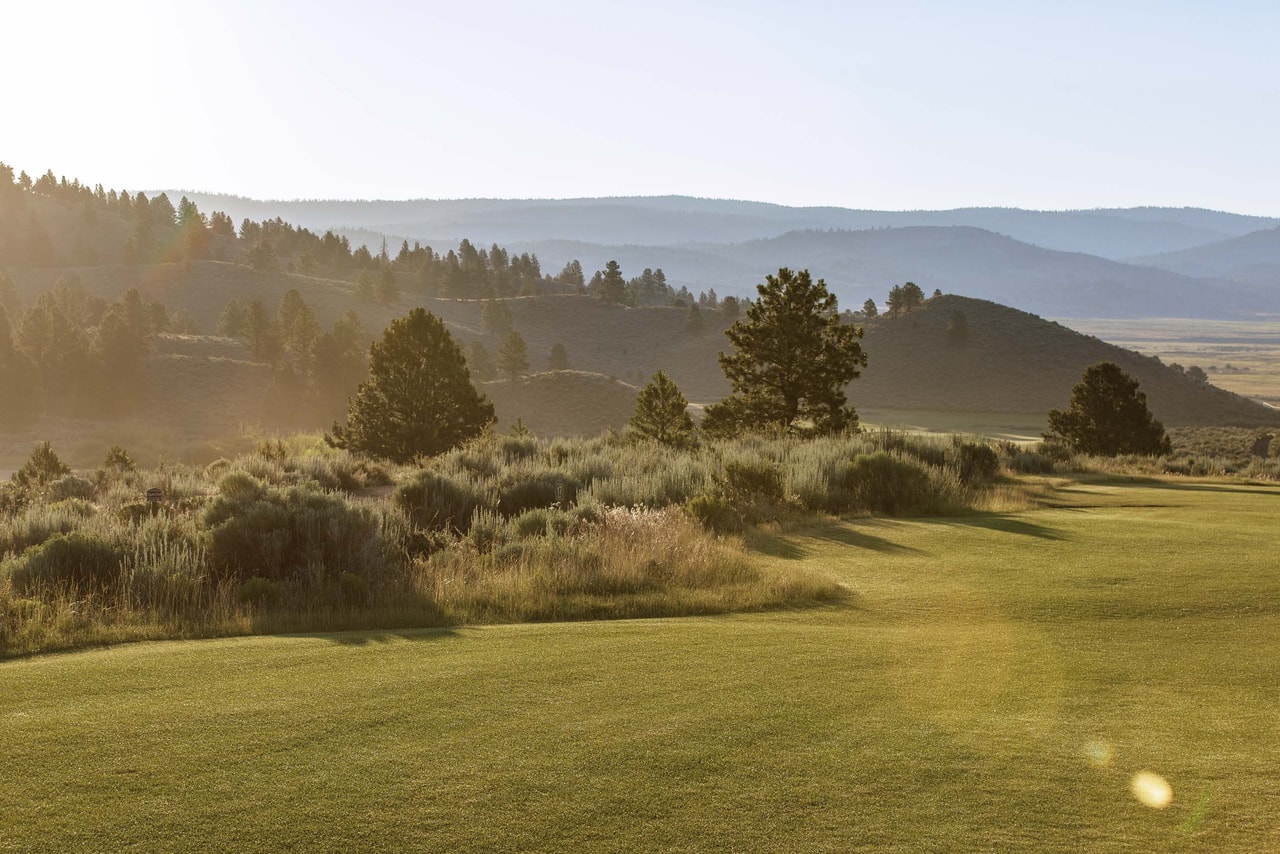 A fairway in hazy low sun, sage and scattered pines along its edge, hills fading into the distance