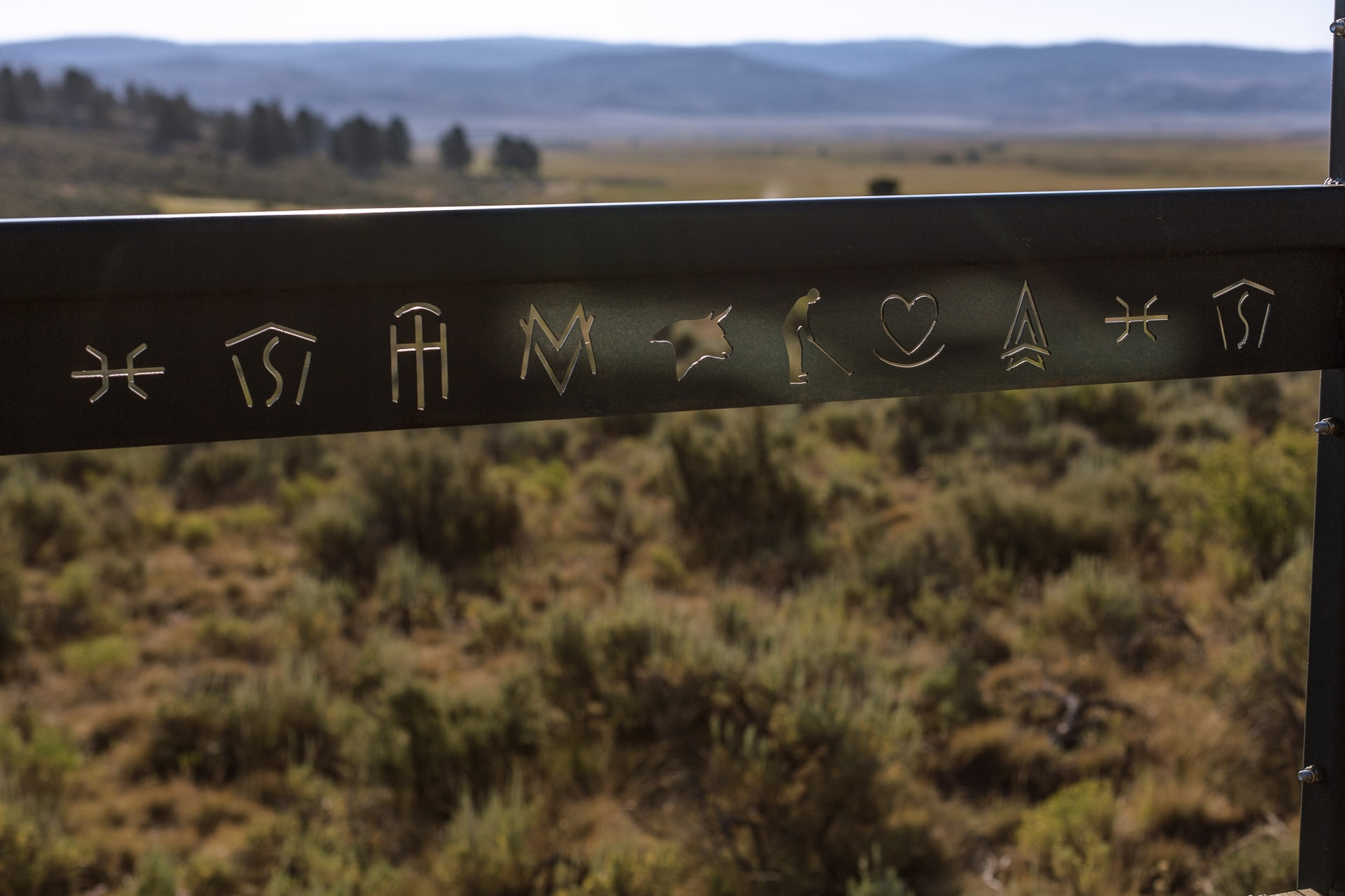 A black metal rail cut with brand-like symbols, a golfer and a heart among them, above sagebrush and hazy hills