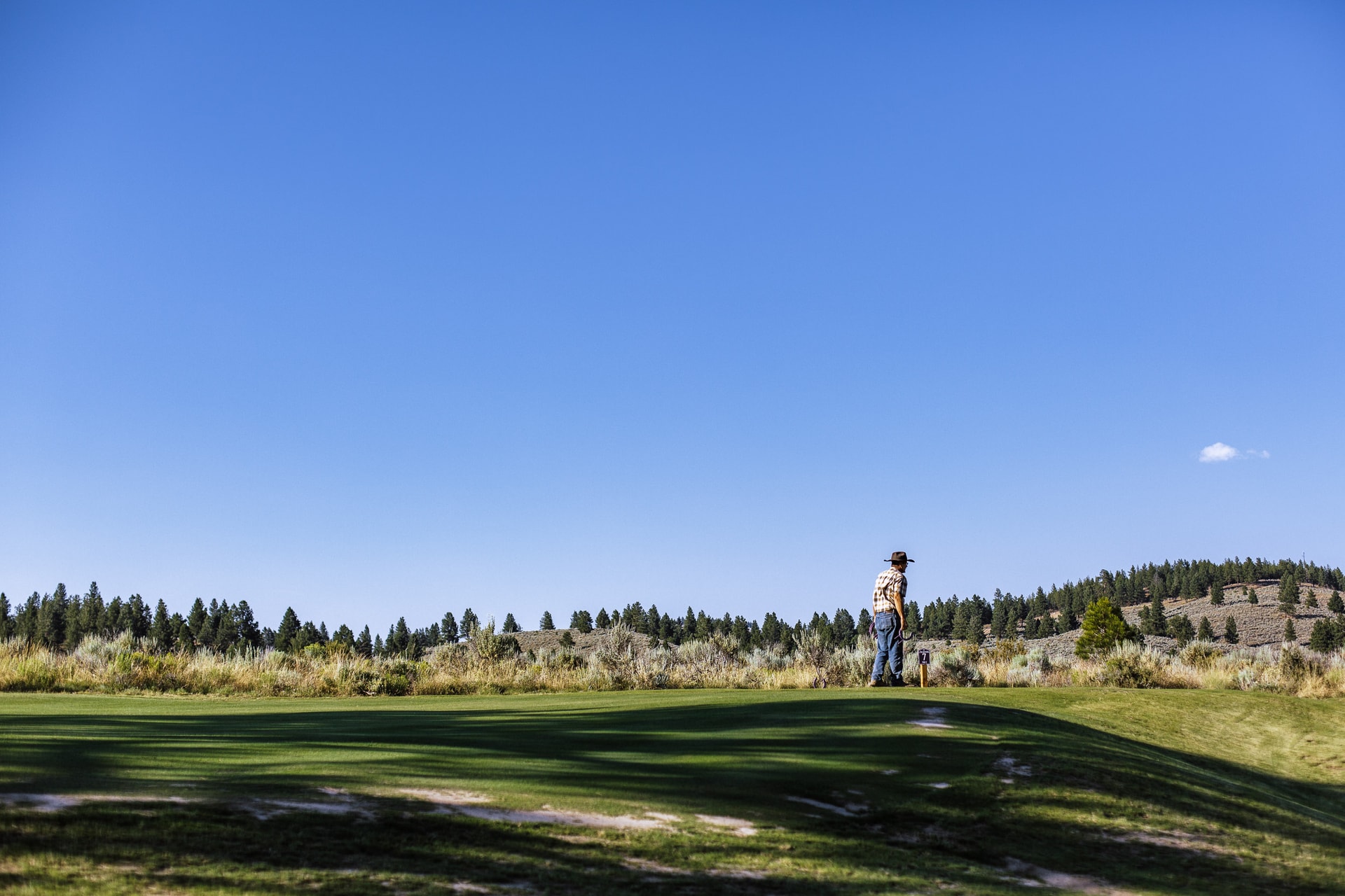 A man in a cowboy hat and checked shirt stands small at the far edge of a fairway beneath a vast blue sky